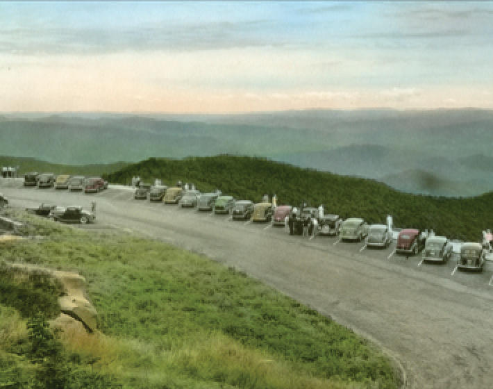 Early 1900s colorized photograph of Great Smoky Mountains featuring line of vehicles parked at a designated pull-off with view of mountain vista
