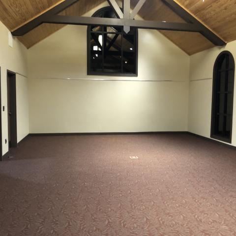 Photo shows a carpeted room with two windows without blinds or curtains. The ceiling is vaulted. Power outlets are in the middle of the floor.