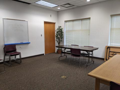 Photo shows a carpeted room, windows with blinds, folding tables and stackable chairs. A dry-erase board is on one wall.