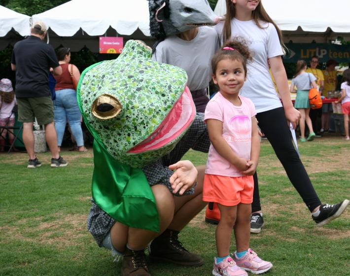 young girl smirks at person in papier-mâché frog costume